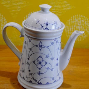 Peut inclure: Une cafetière en céramique blanche avec un motif floral bleu et blanc. La cafetière a un couvercle et un bec.