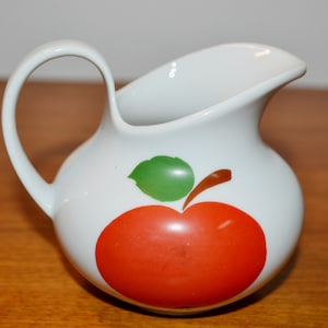 Peut inclure: Une crémier en céramique blanche avec un motif de pomme rouge. La pomme a une feuille verte et une tige brune.