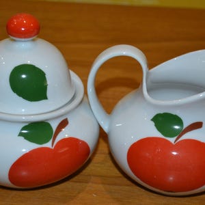 Peut inclure: Un ensemble sucrier et crémier en porcelaine blanche avec des motifs de pommes rouges et de feuilles vertes. Le couvercle du sucrier a un bouton rouge.