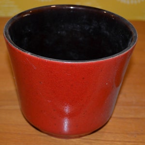 Könnte beinhalten: Ein roter Keramiktopf mit einem schwarzen glasierten Innenraum. Der Topf hat eine abgerundete Form und eine glatte, glänzende Oberfläche.