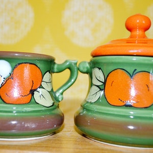 May include: A green ceramic creamer and sugar bowl set with orange lids and painted floral and apple designs.