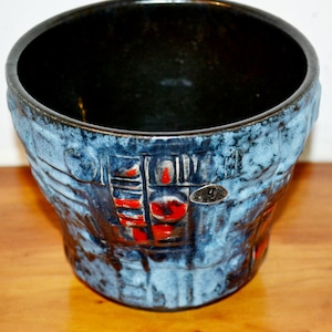 Pode incluir: Um vaso de cerâmica com um exterior azul texturizado e um interior preto. O vaso apresenta detalhes em vermelho e castanho e uma pequena etiqueta prateada. O design é abstrato e geométrico, com um aspeto ligeiramente áspero e feito à mão.