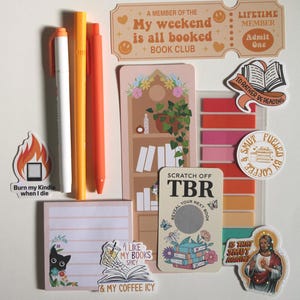 Op de afbeelding: Een verzameling briefpapier en stickers met een boeken thema. Inclusief pennen, plaknotities, een kraslot en stickers met zinnen als "Burn my Kindle when I die" en "My weekend is all booked."
