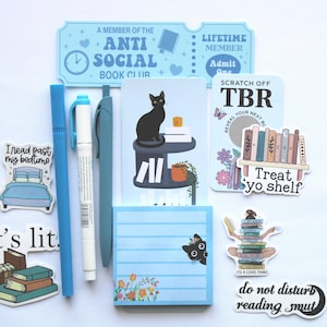 Op de afbeelding: Een verzameling briefpapierartikelen met een boeken thema. Inclusief stickers, pennen, een notitieblok en een ticketvormig item. De stickers bevatten zinnen als "It's lit" en "do not disturb reading smut". Het kleurenschema is voornamelijk blauw en wit.