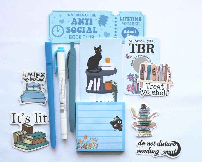 Op de afbeelding: Een verzameling briefpapierartikelen met een boeken thema. Inclusief stickers, pennen, een notitieblok en een ticketvormig item. De stickers bevatten zinnen als "It's lit" en "do not disturb reading smut". Het kleurenschema is voornamelijk blauw en wit.