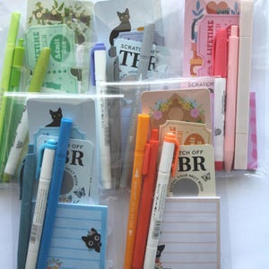 Op de afbeelding: Assortiment van briefpapierartikelen in een doorzichtige plastic verpakking. Inclusief pennen, markeerstiften, notitieblokken en decoratieve kaarten met bloemen- en kattenillustraties. Sommige kaarten hebben de tekst "Admit One" en "Scratch Off".