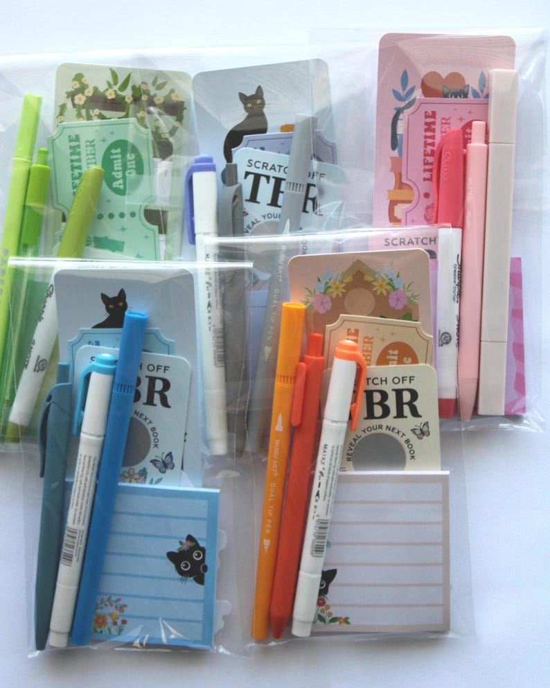 Op de afbeelding: Assortiment van briefpapierartikelen in een doorzichtige plastic verpakking. Inclusief pennen, markeerstiften, notitieblokken en decoratieve kaarten met bloemen- en kattenillustraties. Sommige kaarten hebben de tekst "Admit One" en "Scratch Off".