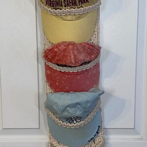 May include: A macrame wall hanging with a gold ring at the top.  Four baseball caps are hanging from the ring. The caps are yellow, red, blue, and white. The yellow cap has the text "VIRGINIA SAFARI PARK" on it. The blue cap has the text "Life is Good" on it.