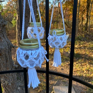 Peut inclure: Trois bougeoirs en crochet blanc avec des couvercles dorés suspendus à une clôture en métal noir. Chaque bougeoir a un pompon blanc.