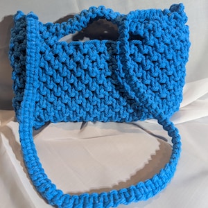 May include: A bright blue, handmade macrame tote bag. The bag features a woven design with two short handles and a long shoulder strap. The bag is made of a soft, textured material.