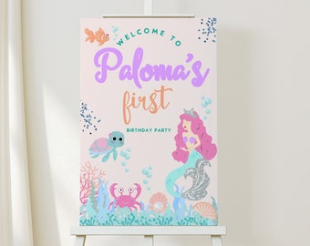 May include: A pink and blue welcome sign for a first birthday party. The sign says "Welcome to Paloma's first birthday party". It features a mermaid with pink hair and a turquoise tail, a sea turtle, a crab, and a fish.
