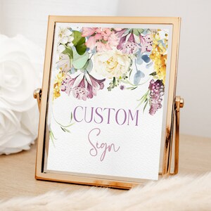 May include: A gold-framed sign with a floral design featuring pink, purple, and yellow flowers. The text "CUSTOM Sign" is written in purple on the sign.