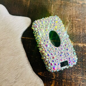 Pu&ograve; includere: Una custodia rettangolare tempestata di strass iridescenti. La custodia ha un'apertura ovale verde scuro e un piccolo display digitale. La custodia &egrave; su una superficie di legno.