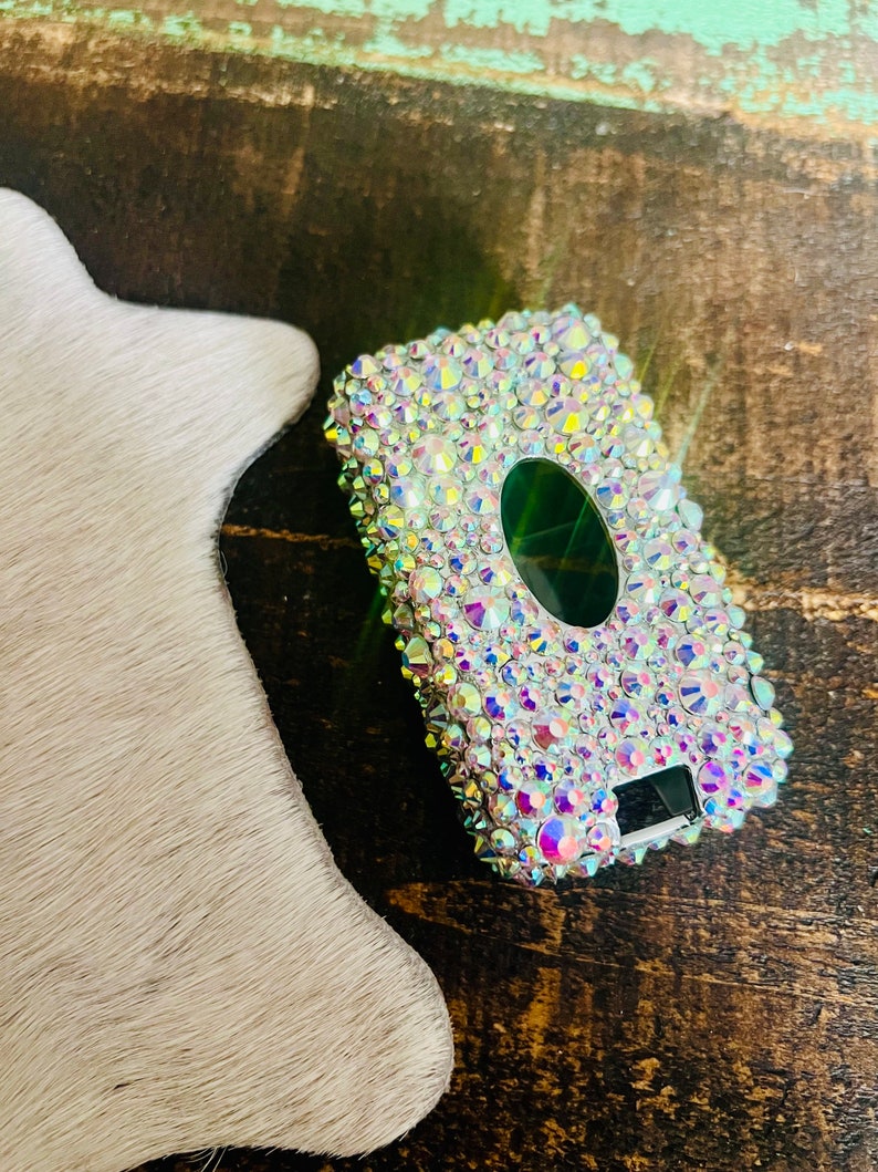 Pu&ograve; includere: Una custodia rettangolare tempestata di strass iridescenti. La custodia ha un'apertura ovale verde scuro e un piccolo display digitale. La custodia &egrave; su una superficie di legno.