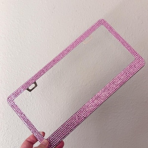 May include: A pink license plate frame covered in small, sparkling rhinestones. The frame is rectangular with rounded corners and a clear center. The frame is held up against a white wall.