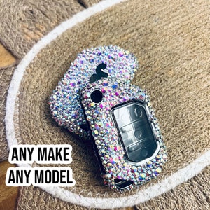 May include: A car key fob covered in iridescent rhinestones. The key fob is silver with a black button and a clear window. The text "ANY MAKE ANY MODEL" is in a white box in the lower left corner.