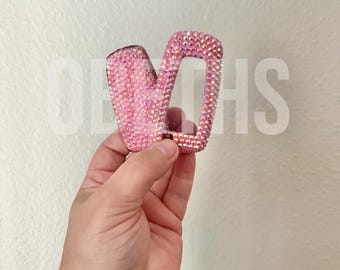 May include: A rectangular hair claw clip covered in small, light pink, reflective rhinestones. The clip features a hinged design with interlocking teeth, creating a sparkling, textured surface that catches the light from multiple angles.