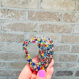 Op de afbeelding: Een met strass versierde, veelkleurige karabijnhaak. De haak is bedekt met een verscheidenheid aan kleurrijke strass-steentjes, waaronder rood, blauw, geel, groen en oranje. De haak wordt vastgehouden in een hand met een lichtpaars gelakte nagel.