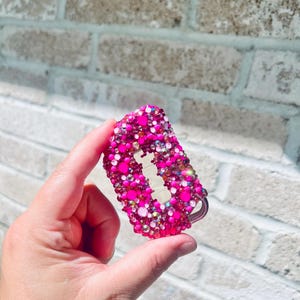 Puede incluir: Un llavero rosa con pedrería y un llavero plateado. El llavero está cubierto de pedrería rosa e iridiscente de varios tamaños. El fondo es una pared de ladrillos de color claro.