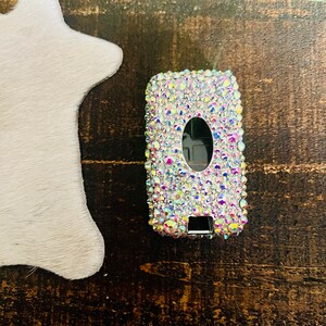Pu&ograve; includere: Una custodia rettangolare per telecomando per auto incastonata con strass iridescenti. Il portachiavi ha un pulsante ovale nero e un pulsante rettangolare nero. La custodia &egrave; su una superficie di legno scuro accanto a un oggetto bianco a forma di stella.