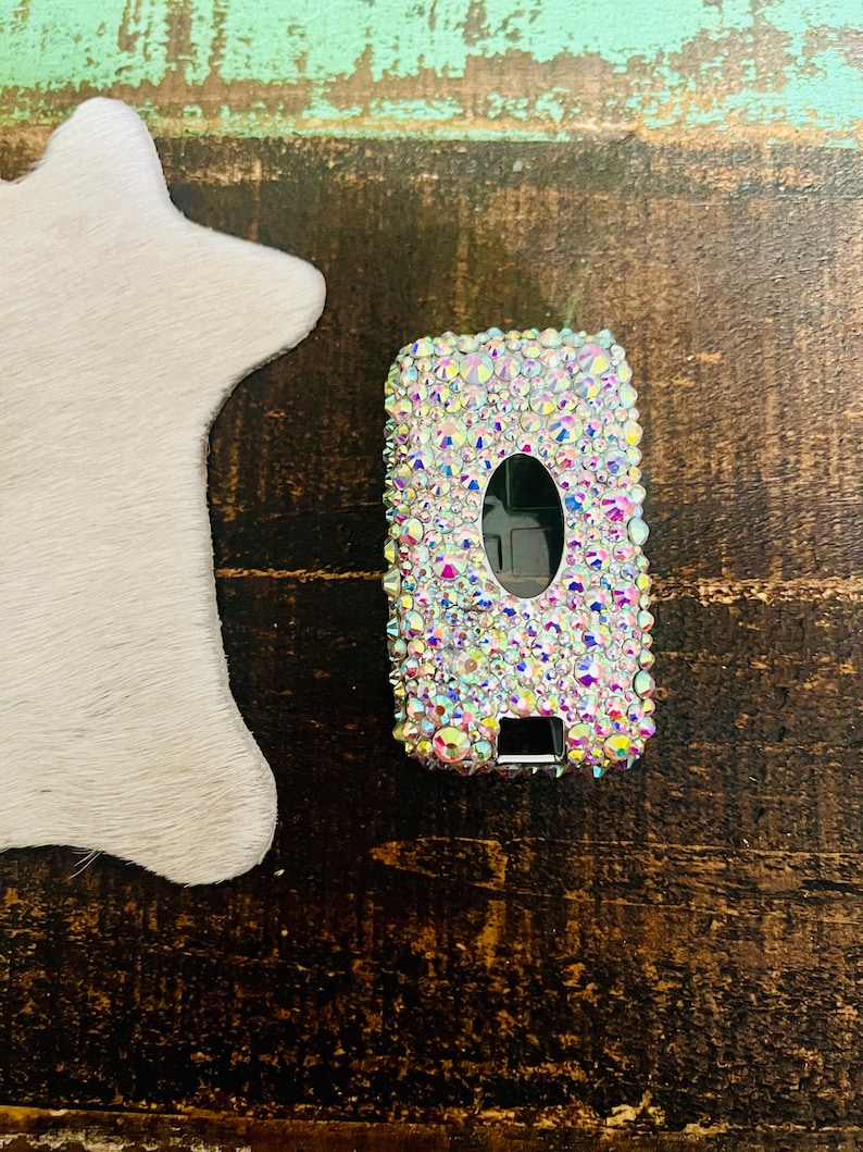 Pu&ograve; includere: Una custodia rettangolare per telecomando per auto incastonata con strass iridescenti. Il portachiavi ha un pulsante ovale nero e un pulsante rettangolare nero. La custodia &egrave; su una superficie di legno scuro accanto a un oggetto bianco a forma di stella.
