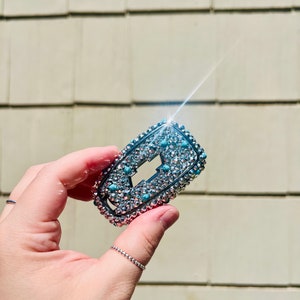 May include: A silver car key fob covered in rhinestones and turquoise blue stones. The key fob is rectangular and has a small opening in the center.