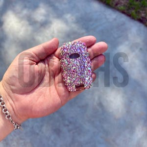 Op de afbeelding: Een autosleutelhanger bedekt met iriserende roze en paarse strass-steentjes. De sleutelhanger heeft een rechthoekige vorm met afgeronde randen en een kleine ovale knop. De strass-steentjes zijn dicht op elkaar gepakt, wat een sprankelend effect creëert.