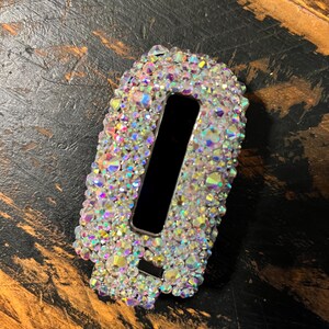 Pu&ograve; includere: Un oggetto rettangolare ricoperto di cristalli iridescenti con un'apertura rettangolare nera. I cristalli riflettono pi&ugrave; colori. L'oggetto &egrave; posto su uno sfondo scuro e strutturato.