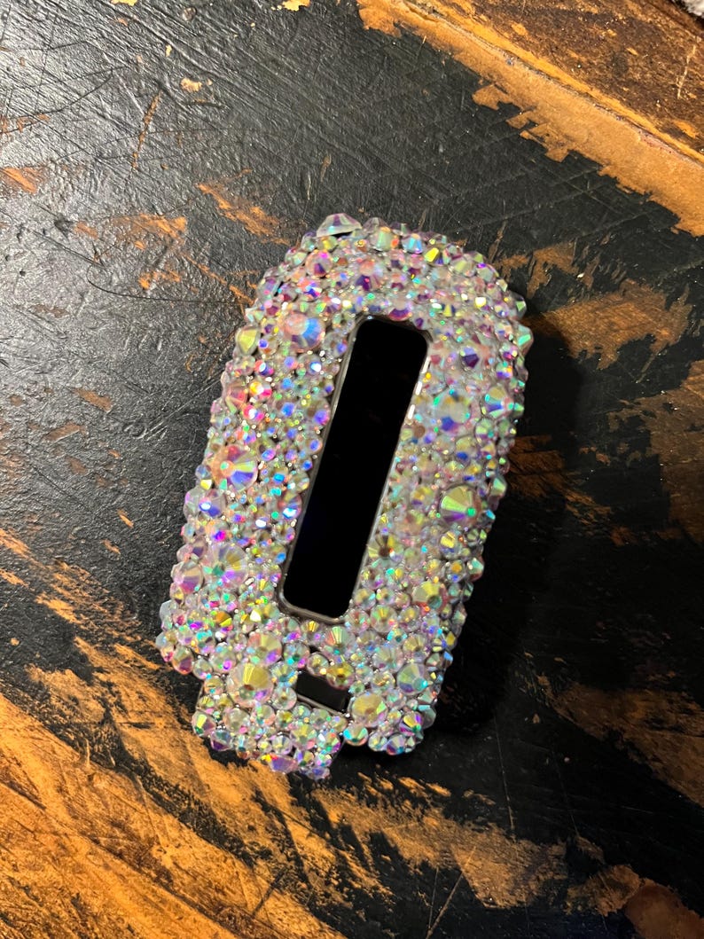Pu&ograve; includere: Un oggetto rettangolare ricoperto di cristalli iridescenti con un'apertura rettangolare nera. I cristalli riflettono pi&ugrave; colori. L'oggetto &egrave; posto su uno sfondo scuro e strutturato.