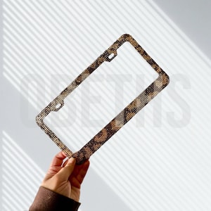 May include: A decorative license plate frame with a brown and tan animal print pattern. The frame is rectangular with rounded corners and has holes for mounting. The frame is held up against a white background with shadows.
