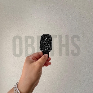 May include: A black, bedazzled microphone-shaped accessory. The accessory is held in a hand, with a silver bracelet visible. The background is a plain white wall with the word "OBOTHS" faintly visible.