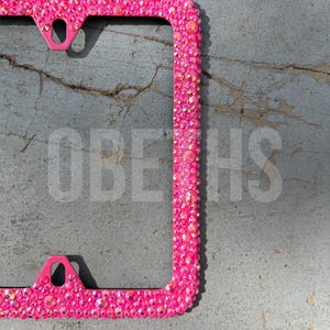 May include: A bright pink license plate frame encrusted with sparkling rhinestones. The frame has a rectangular shape with rounded corners and mounting holes. The background is a textured gray surface.