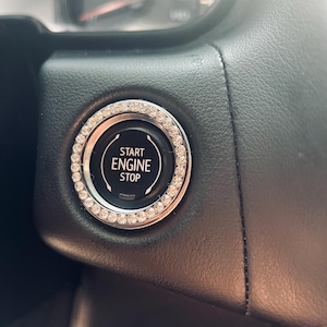 May include: A car's black start/stop button with a silver and rhinestone bezel. The button is surrounded by a black leather dashboard. The text "START ENGINE STOP" is printed on the button.