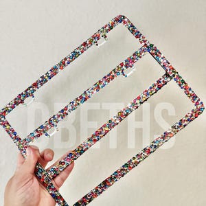 Op de afbeelding: Een rechthoekig kentekenplaathouder volledig bedekt met kleine, veelkleurige strass-steentjes in tinten rood, blauw, groen, geel en transparant. Het frame heeft vier montagegaten en een metalen structuur onder de dichte laag van kristallen.