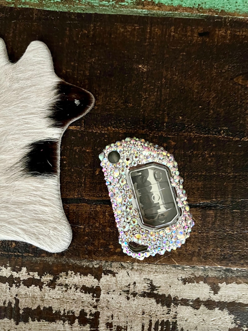 Pu&ograve; includere: Una cover per telecomando auto tempestata di strass iridescenti. Il portachiavi &egrave; argentato e ha pulsanti neri. La cover &egrave; circondata da molti piccoli strass chiari e colorati. Lo sfondo &egrave; una superficie di legno invecchiato.