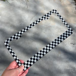 May include: A black and white checkered license plate frame. The frame is decorated with sparkling rhinestones, creating a dazzling effect. The design features a classic checkerboard pattern, adding a touch of glamour to any vehicle.
