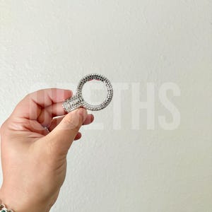 May include: A silver-colored ring with a large, round center stone and smaller stones encircling the band. The ring is held up against a white background. The ring is likely a fashion accessory or a piece of jewelry.