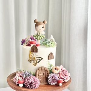May include: A white cake decorated with a fondant fairy figurine, butterflies, flowers, and a small wooden door. The cake is on a wooden cake stand.