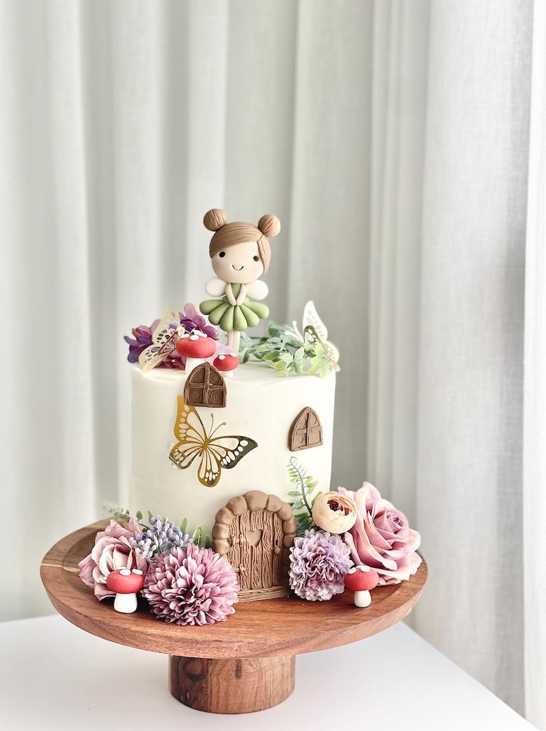 May include: A white cake decorated with a fondant fairy figurine, butterflies, flowers, and a small wooden door. The cake is on a wooden cake stand.