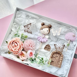 May include: A set of fairy garden cake toppers featuring a pink and purple fairy, a brown door with a heart-shaped window, a pink mushroom, and two butterflies. The toppers are surrounded by pink flowers and green leaves.