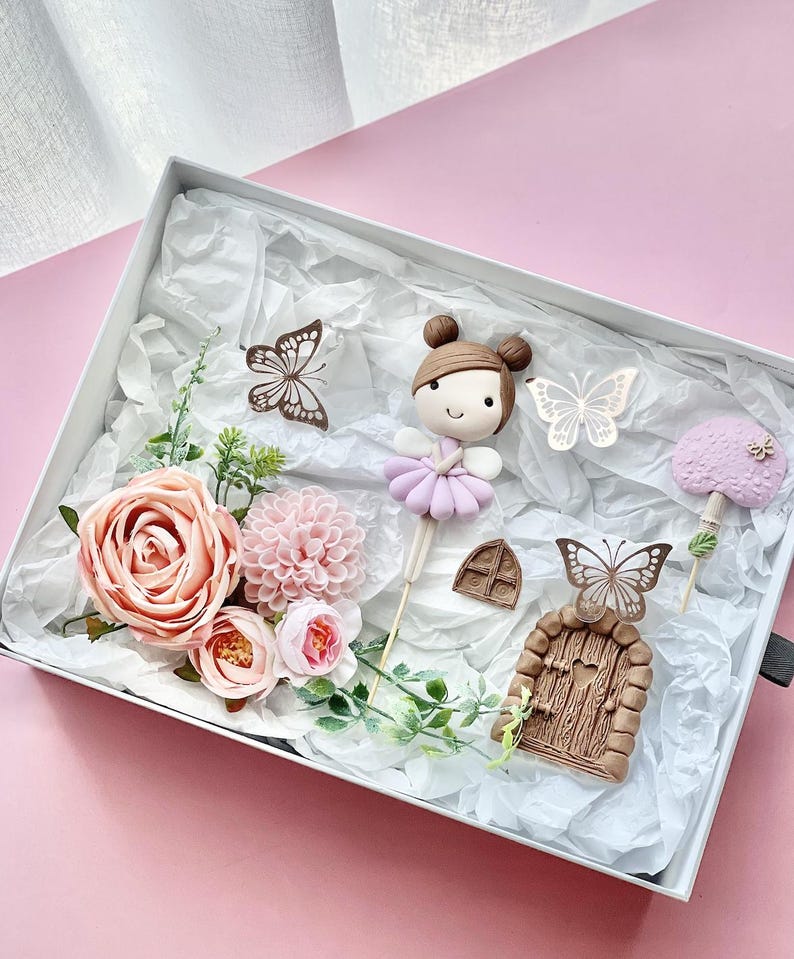 May include: A set of fairy garden cake toppers featuring a pink and purple fairy, a brown door with a heart-shaped window, a pink mushroom, and two butterflies. The toppers are surrounded by pink flowers and green leaves.