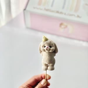May include: A beige, textured bunny figurine topper on a wooden stick. The bunny has a small, light yellow party hat with a circular pattern on its head, dark eyes, and a pink nose. The figure is standing upright with its arms behind its back.