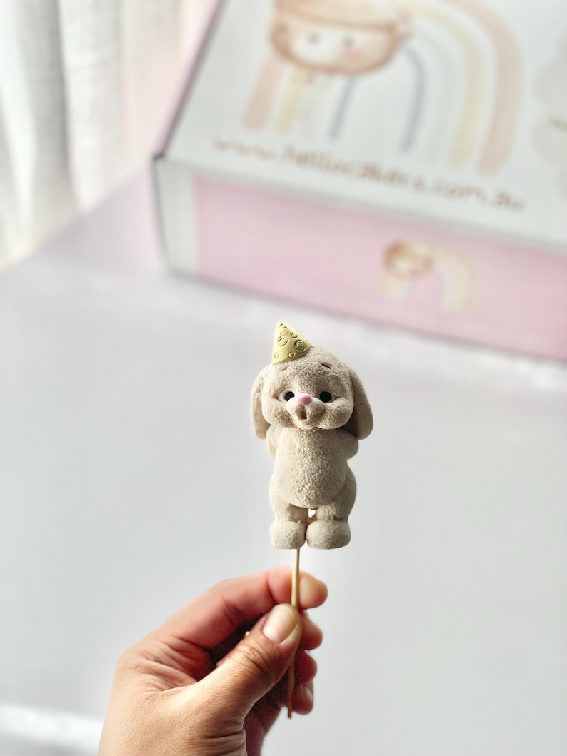 May include: A beige, textured bunny figurine topper on a wooden stick. The bunny has a small, light yellow party hat with a circular pattern on its head, dark eyes, and a pink nose. The figure is standing upright with its arms behind its back.