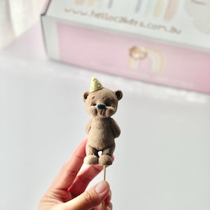 May include: A small, brown, textured bear figurine topper with a light yellow party hat, mounted on a thin wooden stick. The bear has a rounded snout, dark eyes, and a small dark nose, standing upright with its arms positioned at its sides.