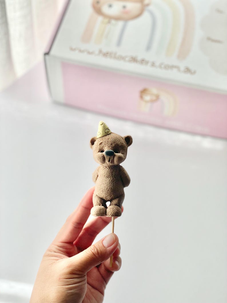 May include: A small, brown, textured bear figurine topper with a light yellow party hat, mounted on a thin wooden stick. The bear has a rounded snout, dark eyes, and a small dark nose, standing upright with its arms positioned at its sides.