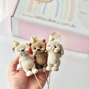 May include: Three small animal cake toppers on wooden sticks, including two bears and one rabbit, each wearing a textured gold party hat. The figures have a matte, granular finish and are positioned in front of a box labeled www.hellocakers.com.au.