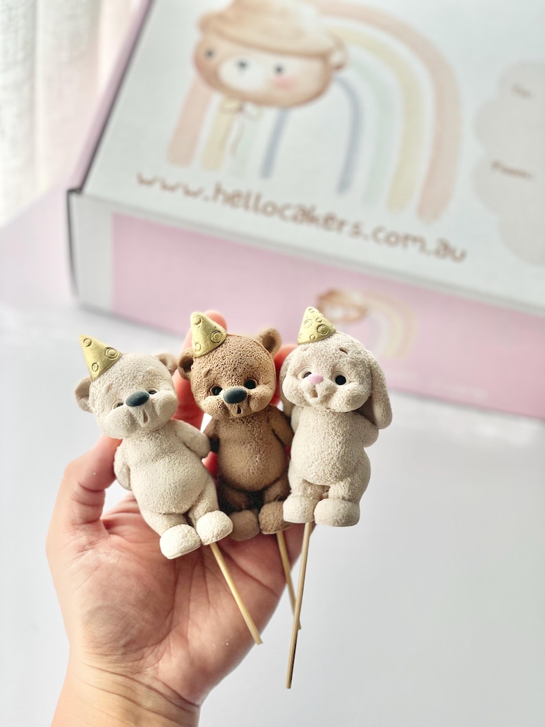 May include: Three small animal cake toppers on wooden sticks, including two bears and one rabbit, each wearing a textured gold party hat. The figures have a matte, granular finish and are positioned in front of a box labeled www.hellocakers.com.au.