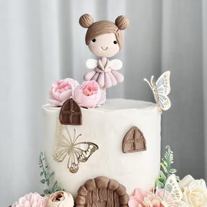 May include: A white cake decorated with pink flowers, butterflies, and a fondant fairy figurine. The cake has a brown door and window details.