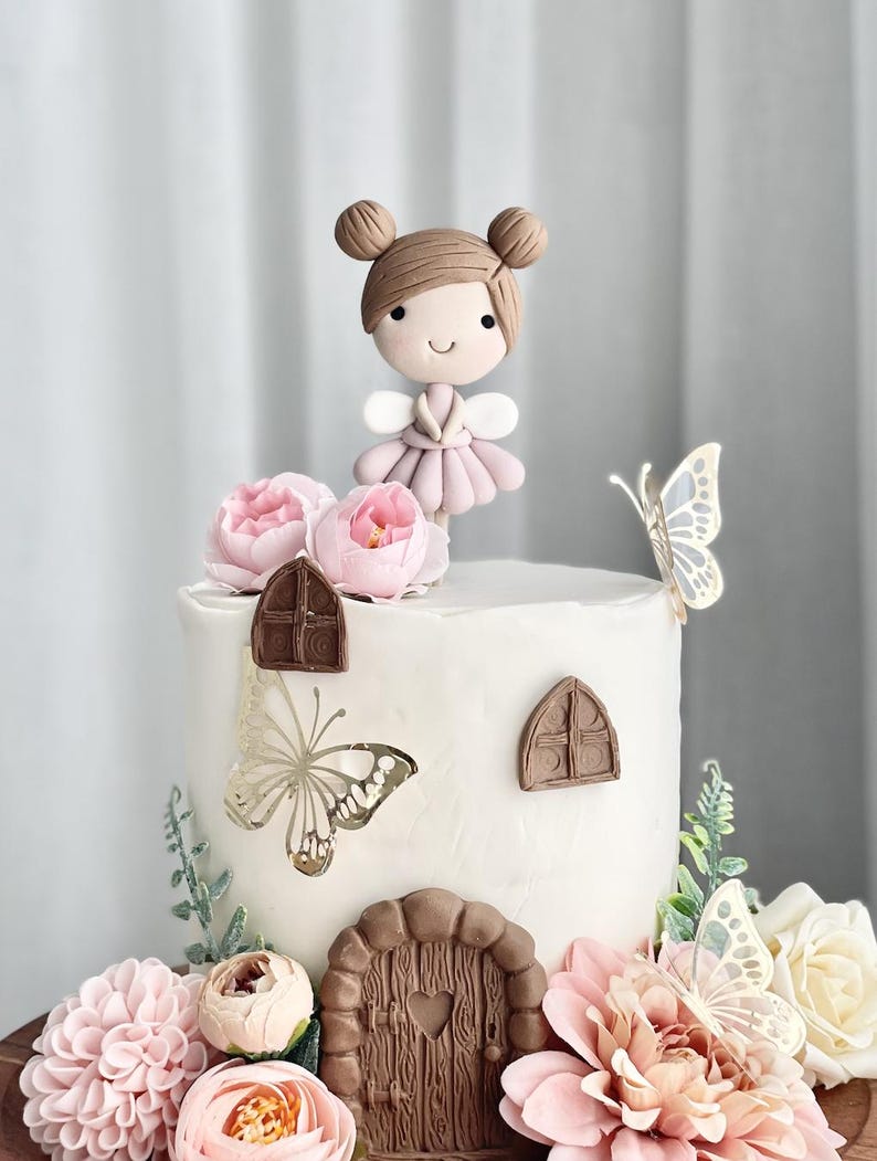 May include: A white cake decorated with pink flowers, butterflies, and a fondant fairy figurine. The cake has a brown door and window details.
