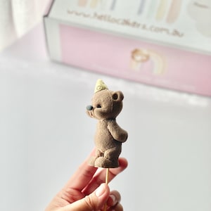 May include: A brown teddy bear cake topper with a textured surface, wearing a small yellow party hat on its head. The bear has a dark nose and is attached to a thin wooden stick for placement. The figure is shown in profile against a blurred box.